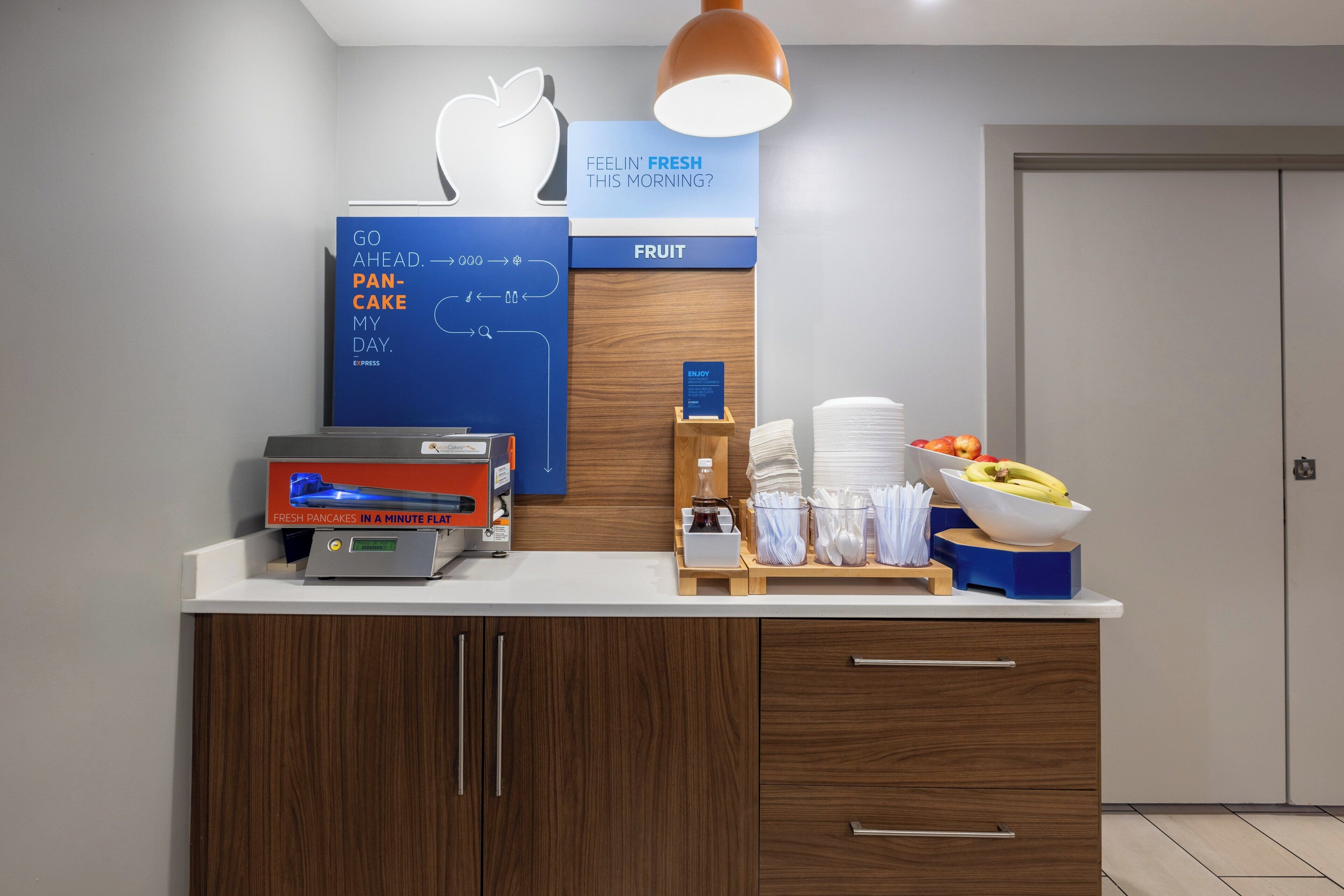 A breakfast station with a sign for fresh pancakes, a fruit display, and a coffee machine.