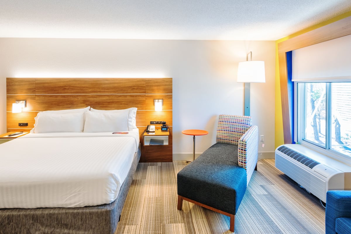 A hotel room with a large bed, a small table, a chair, a window, and an air conditioning unit.
