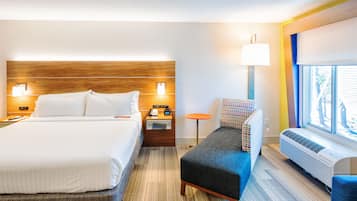 A hotel room with a large bed, a small table, a chair, a window, and an air conditioning unit.