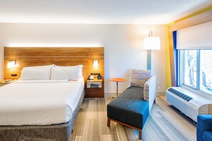 A hotel room with a large bed, a small table, a chair, a window, and an air conditioning unit.