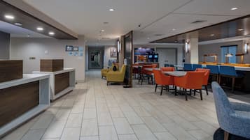 A modern hotel lobby with a reception desk, seating area with orange and blue chairs, and a coffee station.