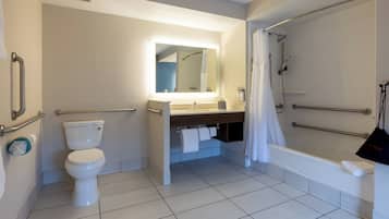 Baño con inodoro, lavamanos, espejo, ducha y barras de apoyo.