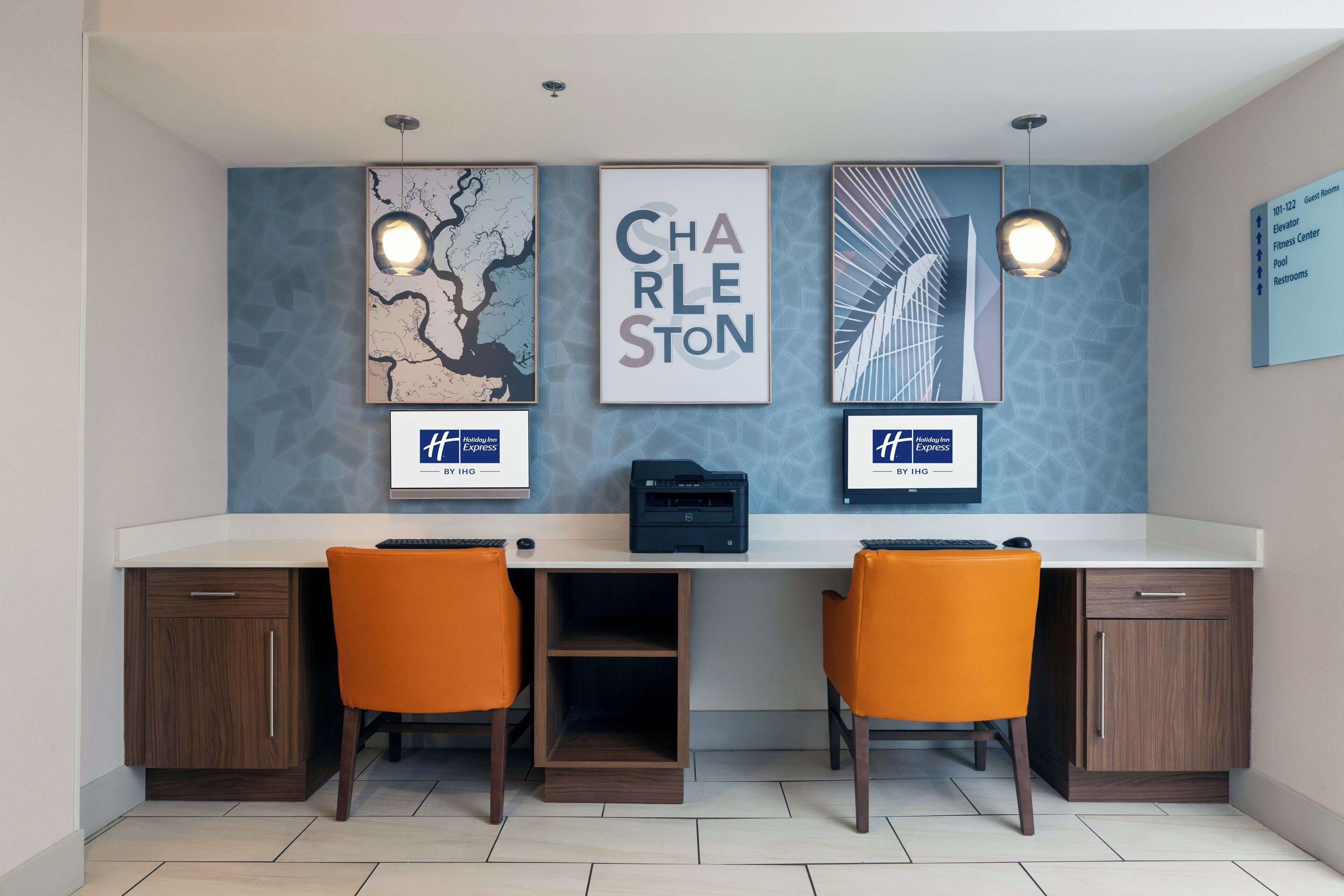 A hotel reception desk with two orange chairs, a printer, framed artwork, and a wall sign indicating amenities.