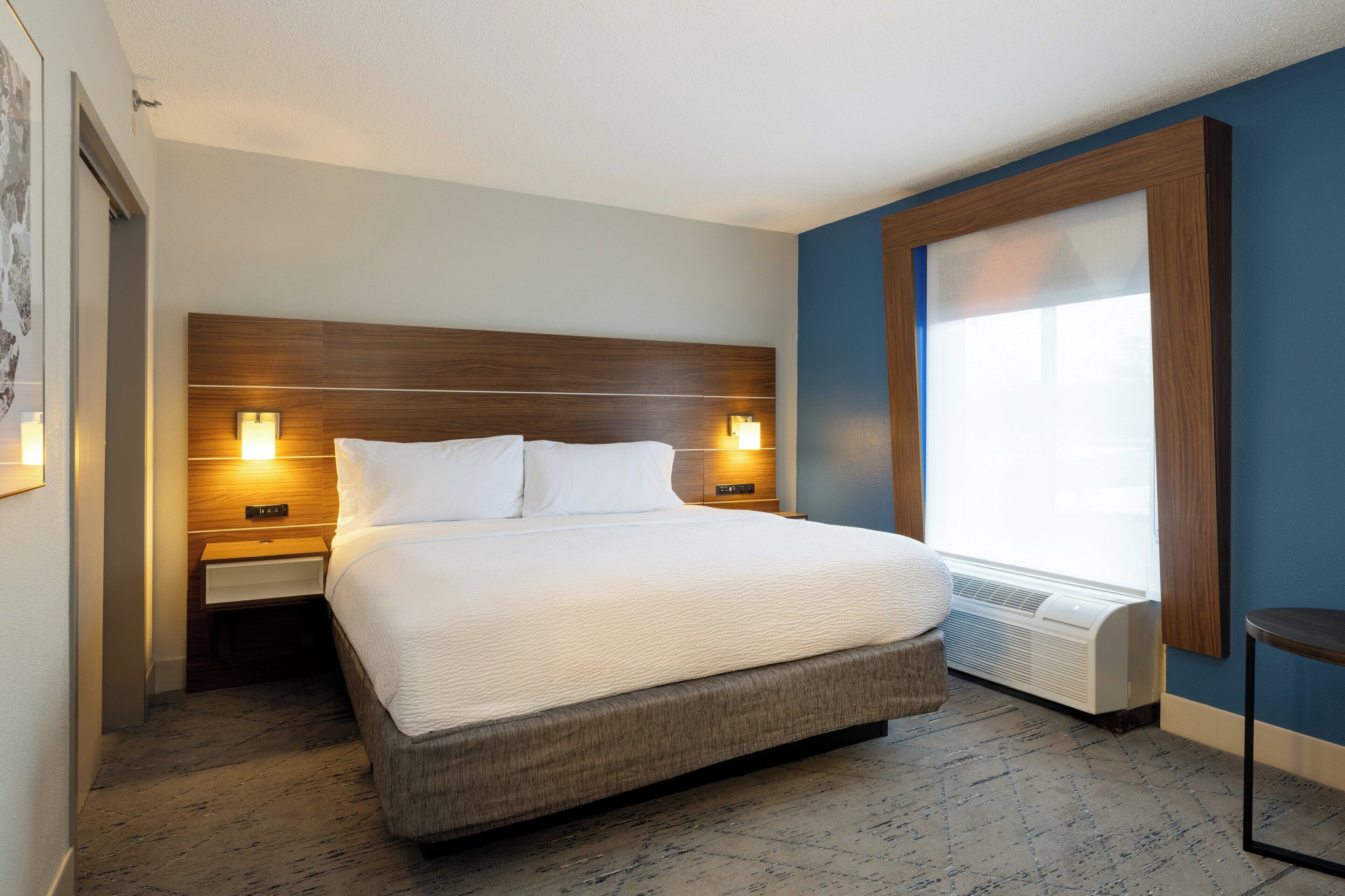 A hotel room with a large bed, wooden headboard, two bedside tables with lamps, a window with blinds, and a wall-mounted air conditioning unit.