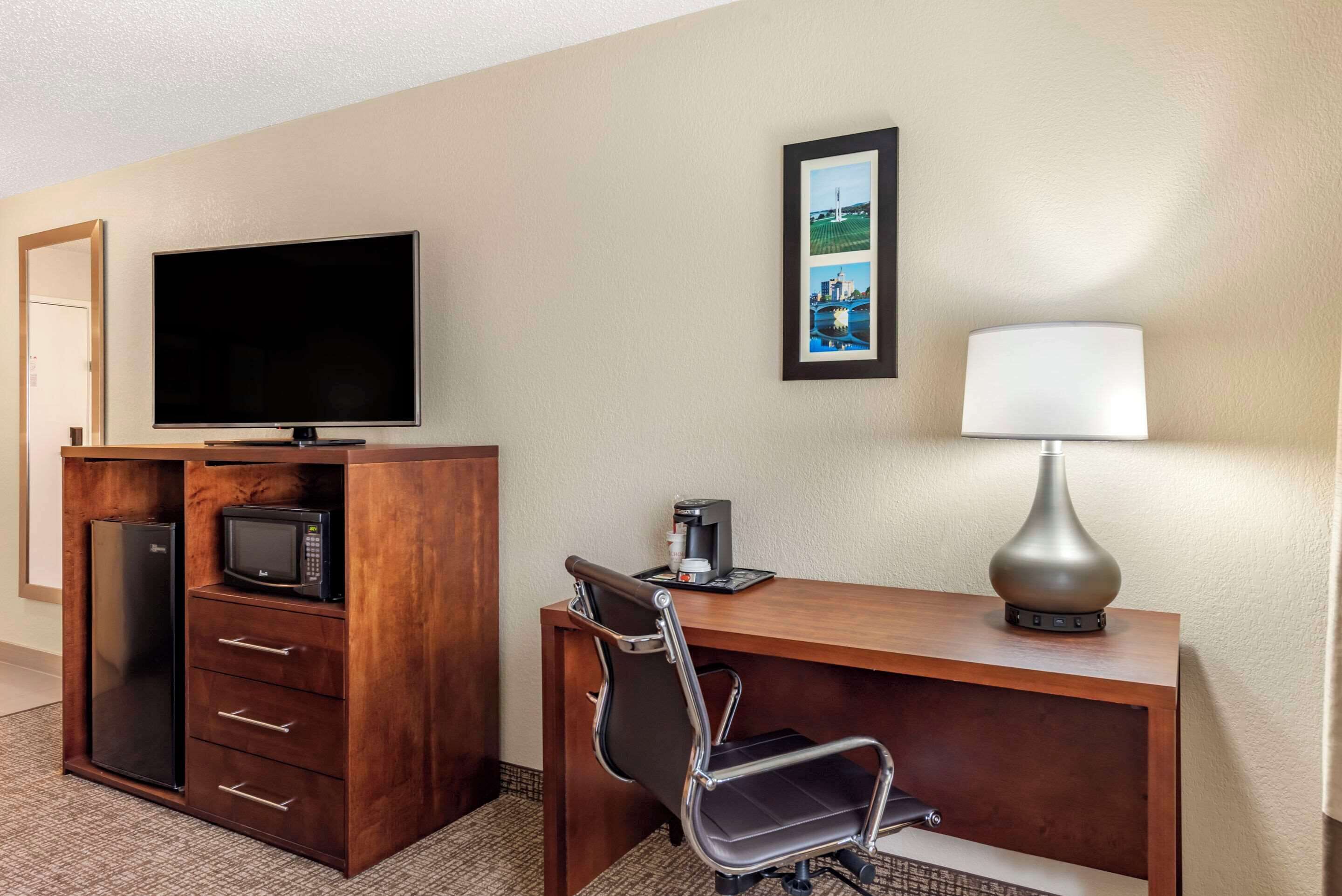A hotel room with a wooden desk, a flat-screen TV, a microwave, a coffee maker, a chair, and a lamp.