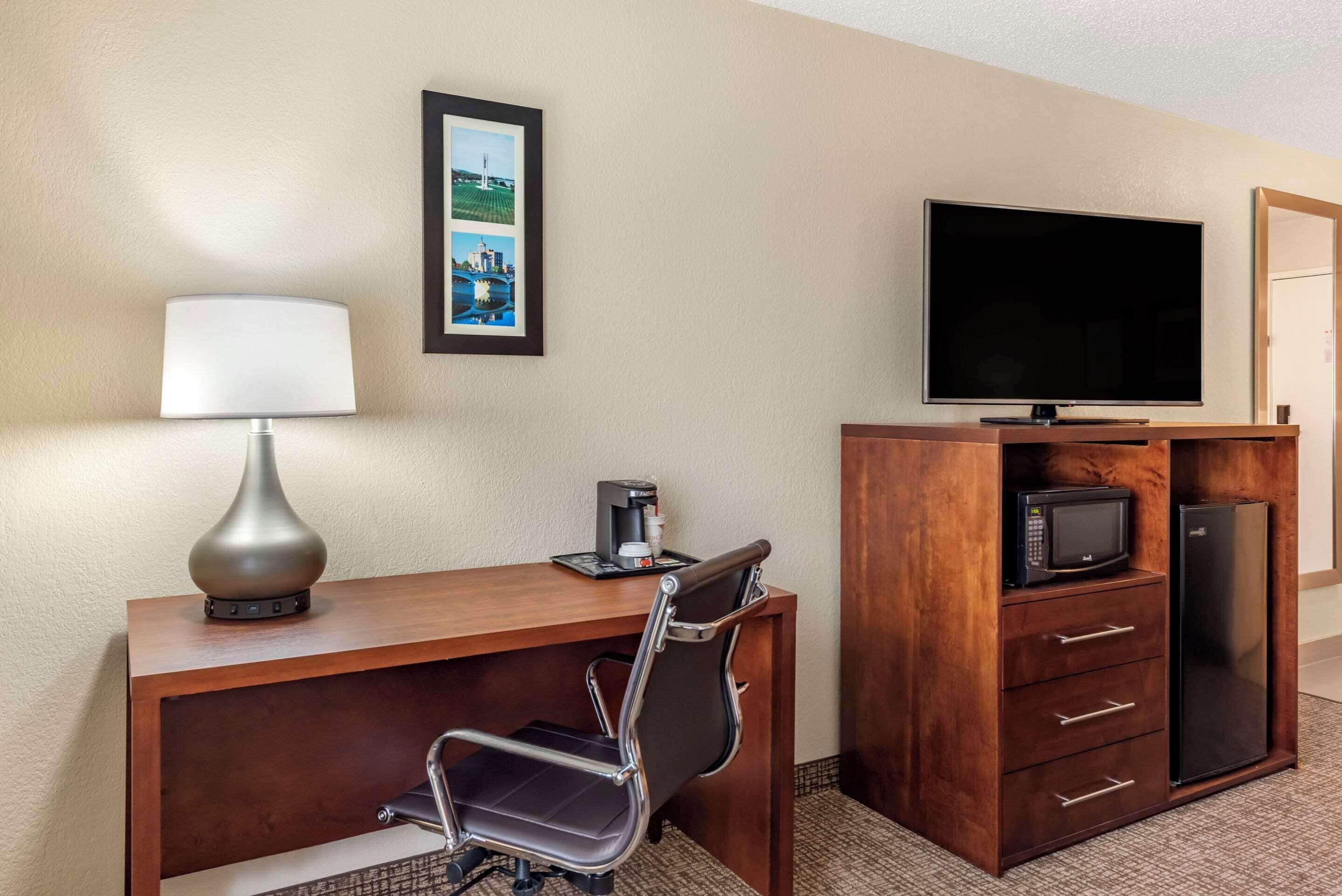 A hotel room with a wooden desk, a chair, a television, a microwave, a refrigerator, a coffee maker, and a framed picture on the wall.
