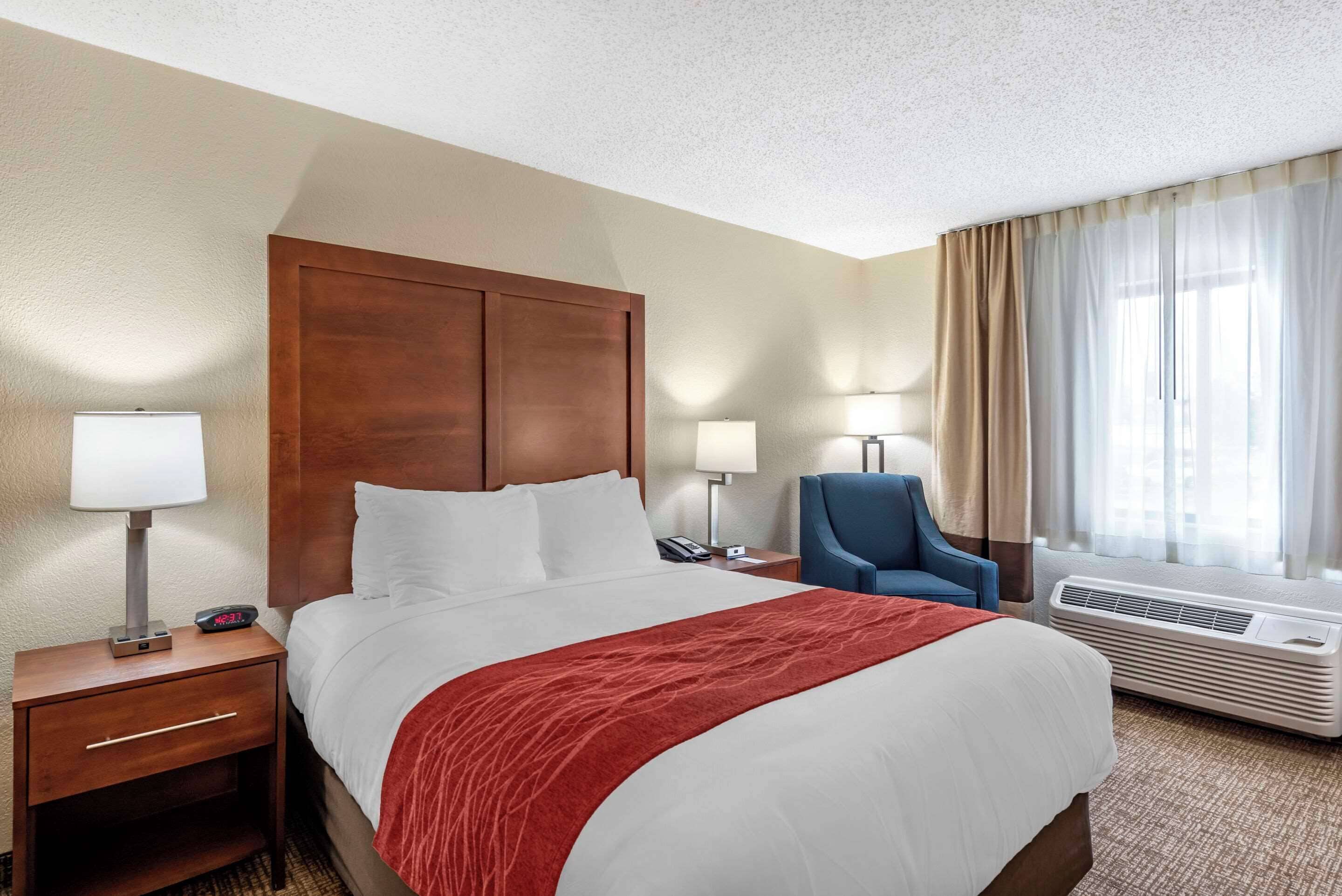 A hotel room with a large bed, a wooden headboard, a desk with a lamp, a chair, and a window with curtains.