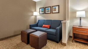 A hotel room with a blue sofa, two matching ottomans, a wooden nightstand, and a lamp. - Comfort Inn & Suites Middletown - Franklin (Franklin)