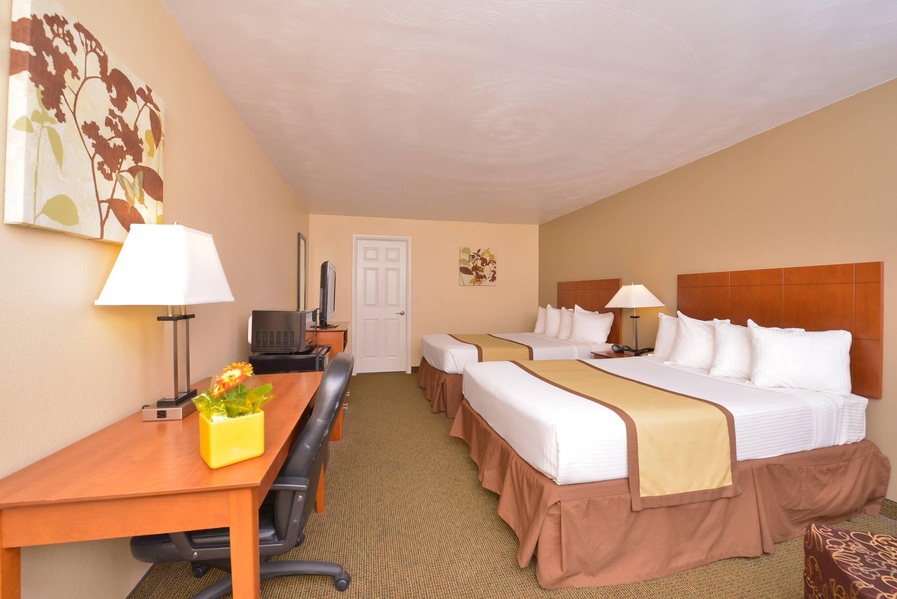 A hotel room with two beds, a desk, a chair, a lamp, and a small table with a flower vase.