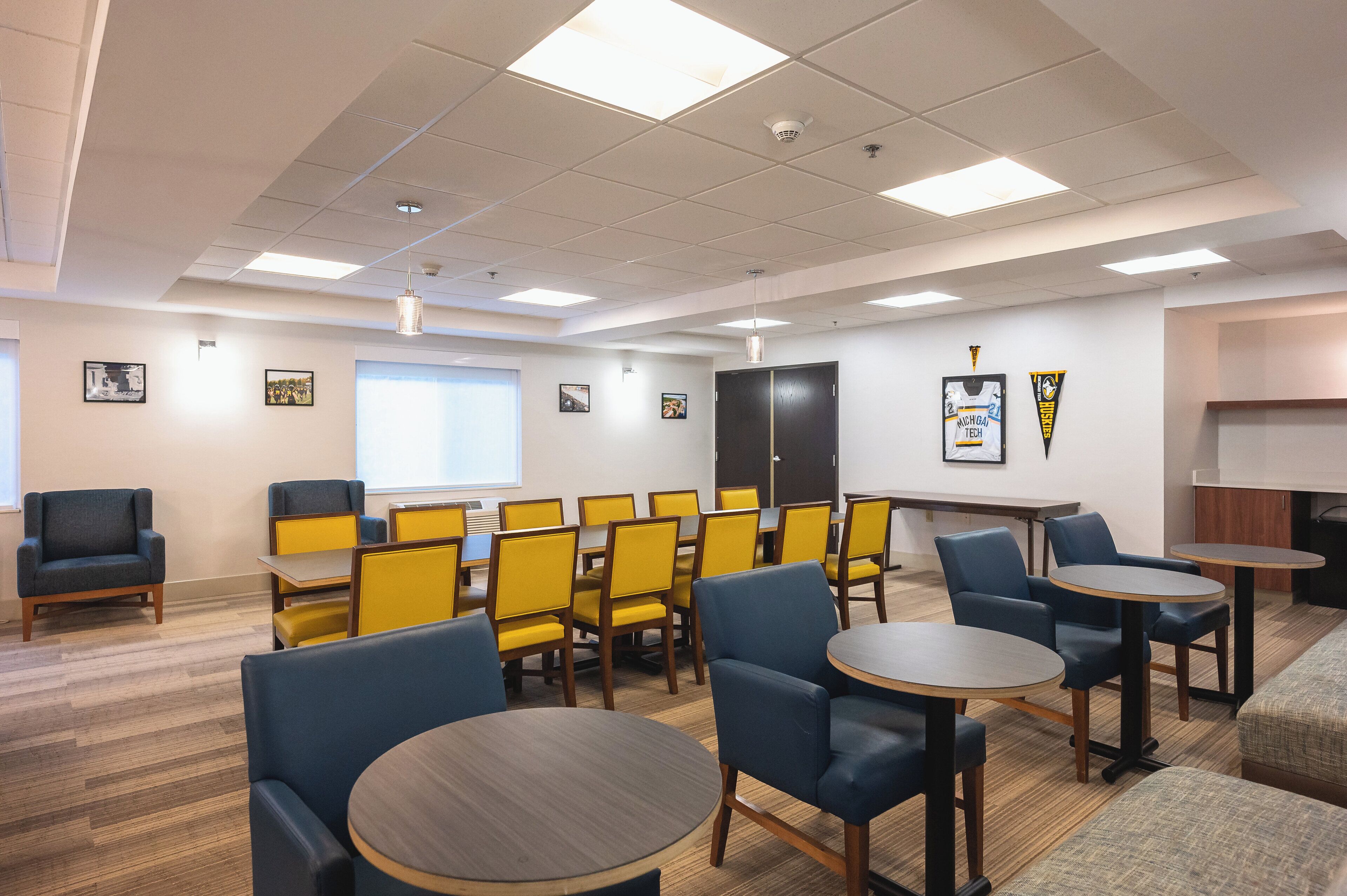 A conference room with yellow and blue chairs, round tables, and a large black cabinet.