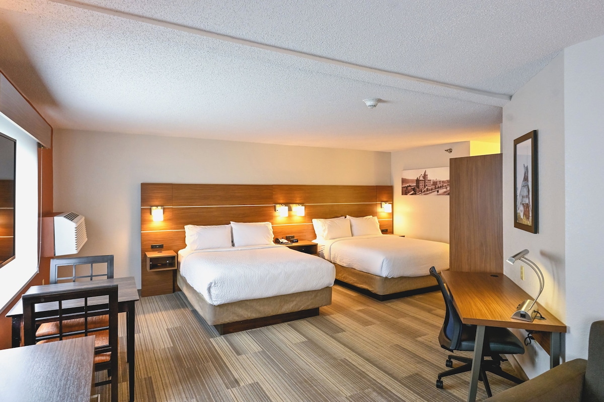 Habitación de hotel con dos camas, un escritorio, una silla, una lámpara y un cuadro en la pared.