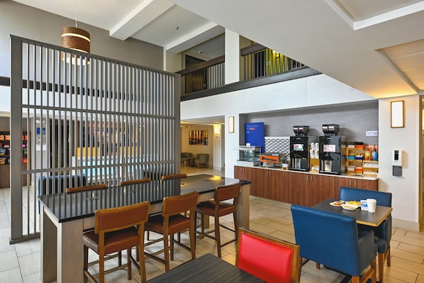 Un moderno vestíbulo de hotel con comedor, estación de café y una escalera que conduce a un nivel superior.