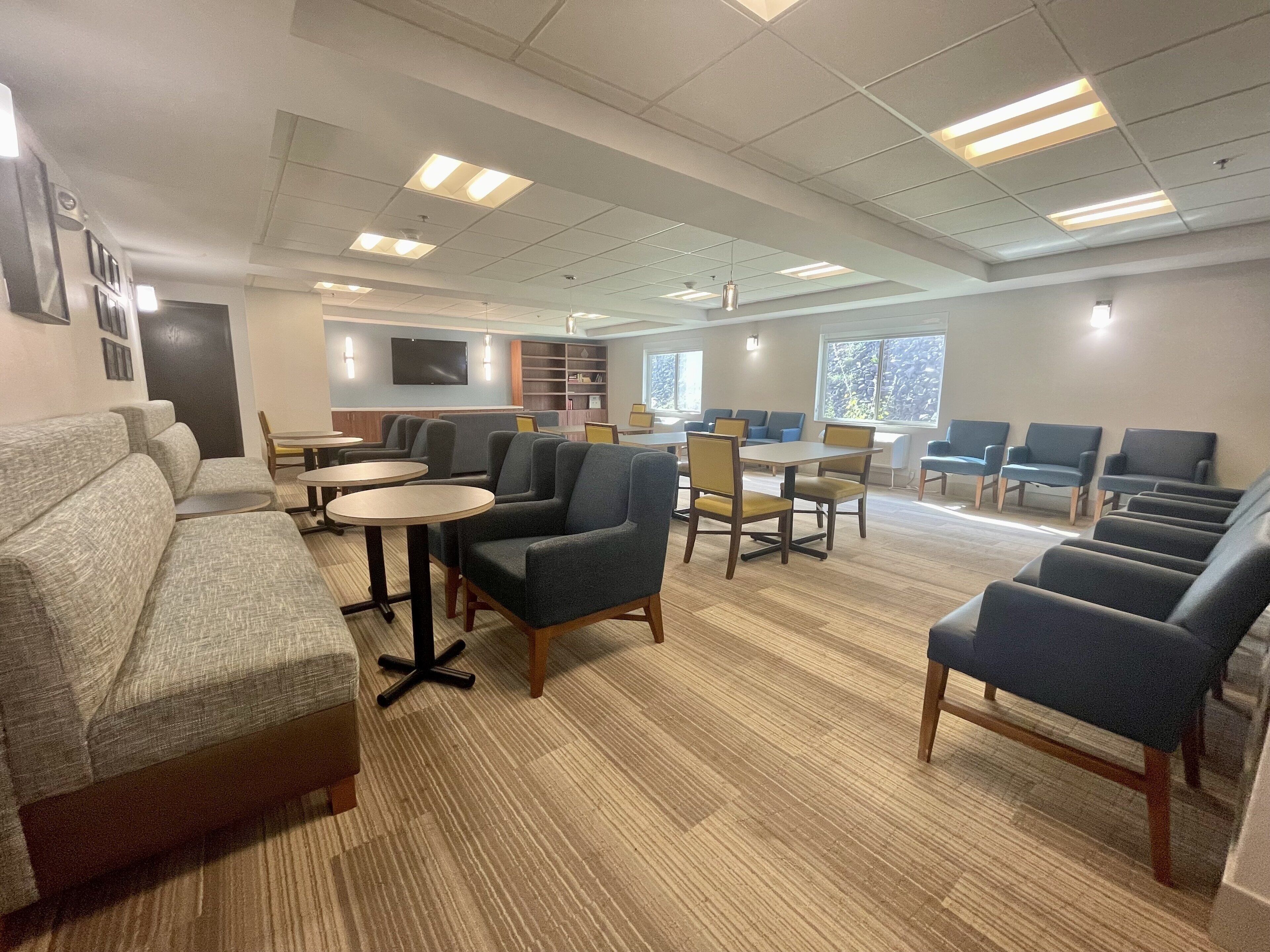 A spacious lounge area with sofas, chairs, and tables, featuring a flat-screen TV and a bookshelf.