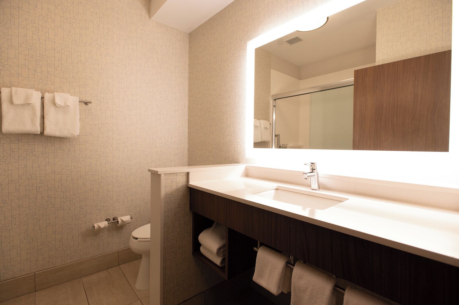 A modern bathroom with a large mirror, a sink, and a toilet. There are towels hanging on the wall and a towel rack.