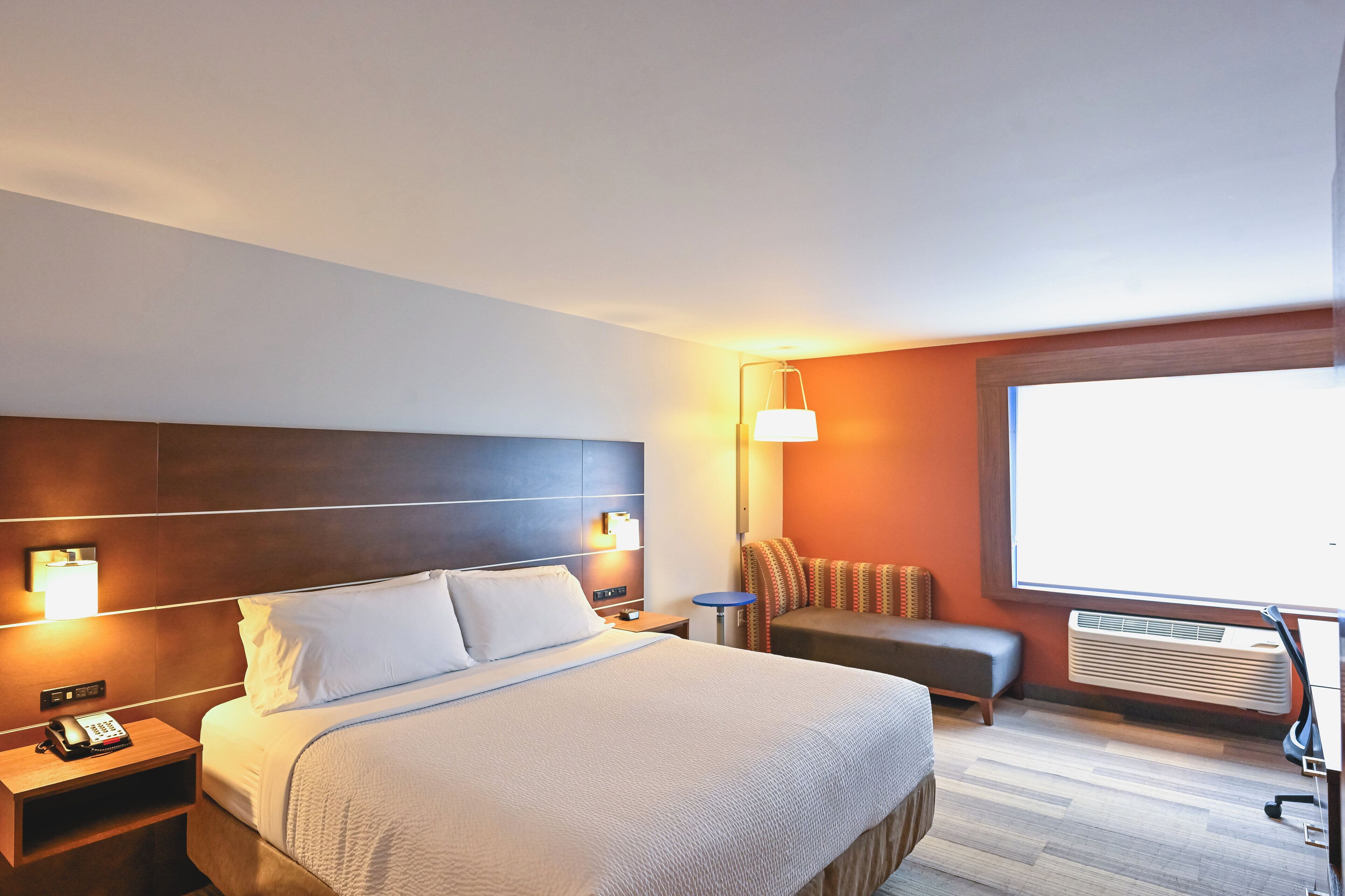 A hotel room with a large bed, bedside tables, a desk, a chair, a sofa, and a large window.