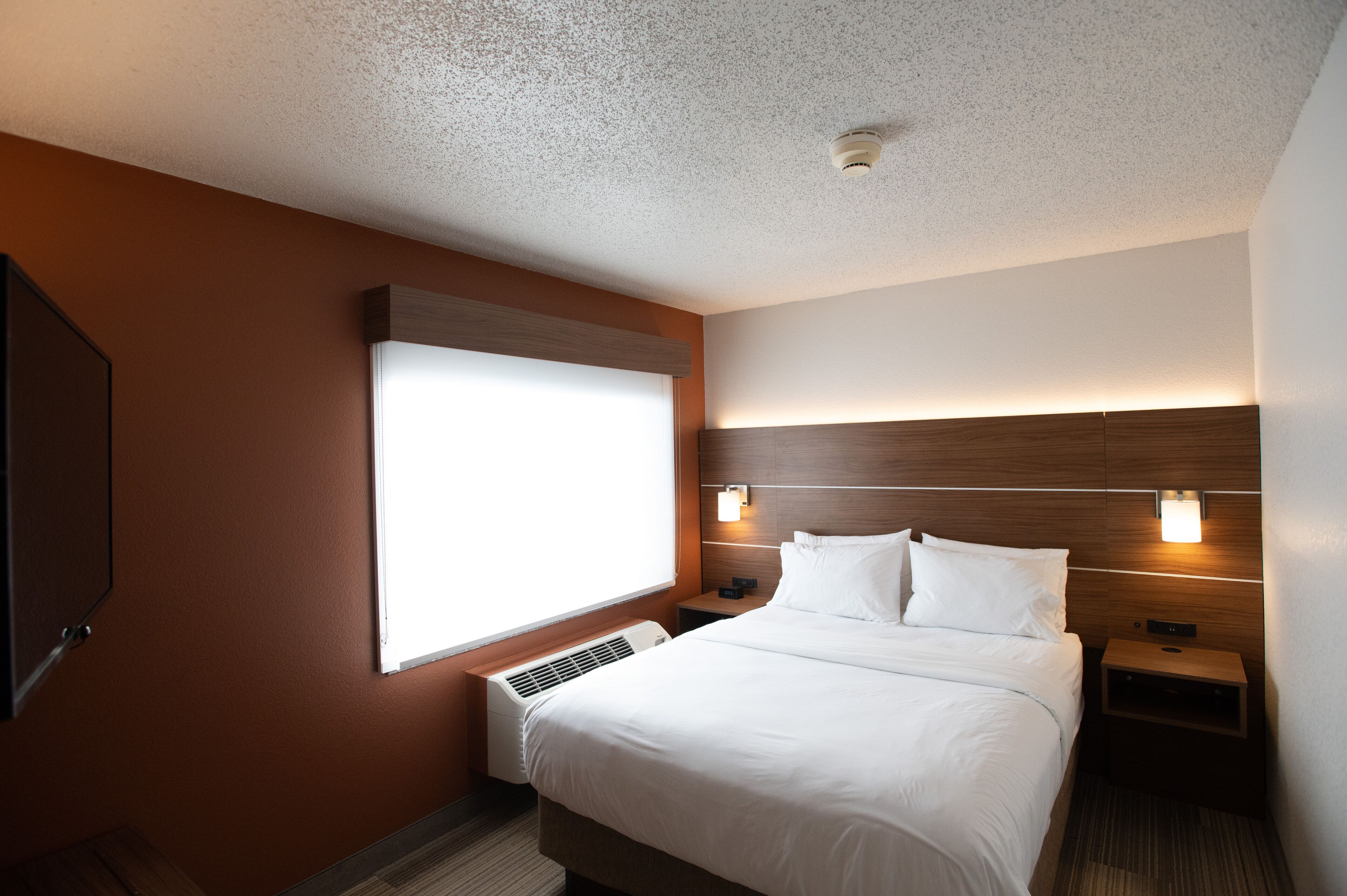A hotel room with a large bed, a wall-mounted TV, a window with blinds, a nightstand, and a wall-mounted light fixture.