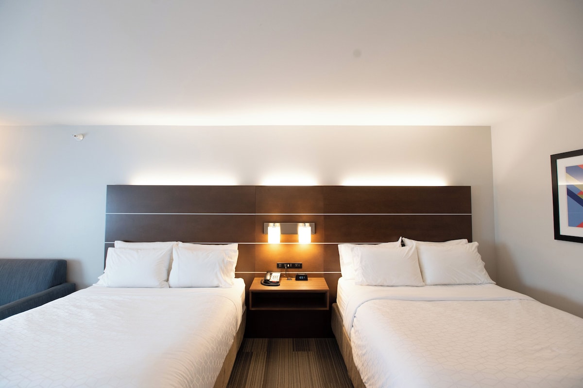 Habitación de hotel con dos camas, una mesita de noche con teléfono y despertador, y una lámpara fijada en la pared.