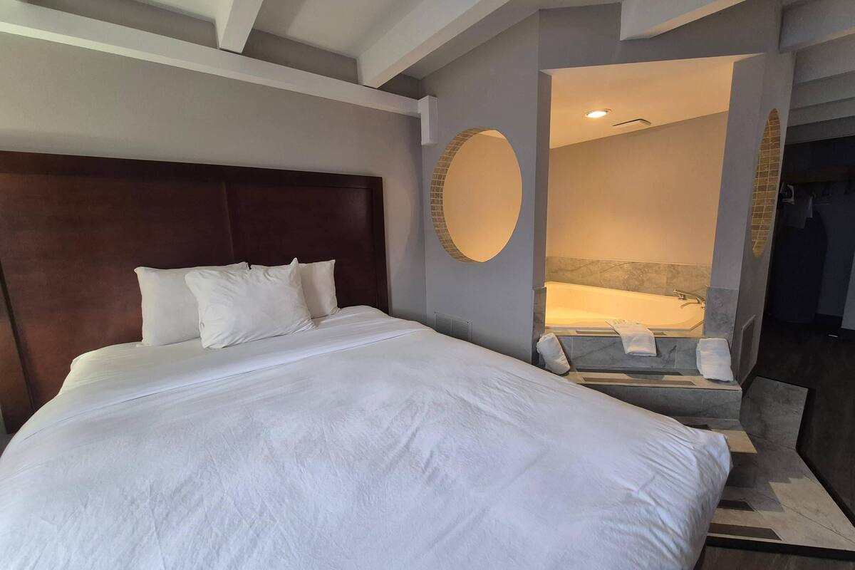 Quarto de hotel moderno com uma cama grande, um espelho redondo e um espaço de banheiro separado.