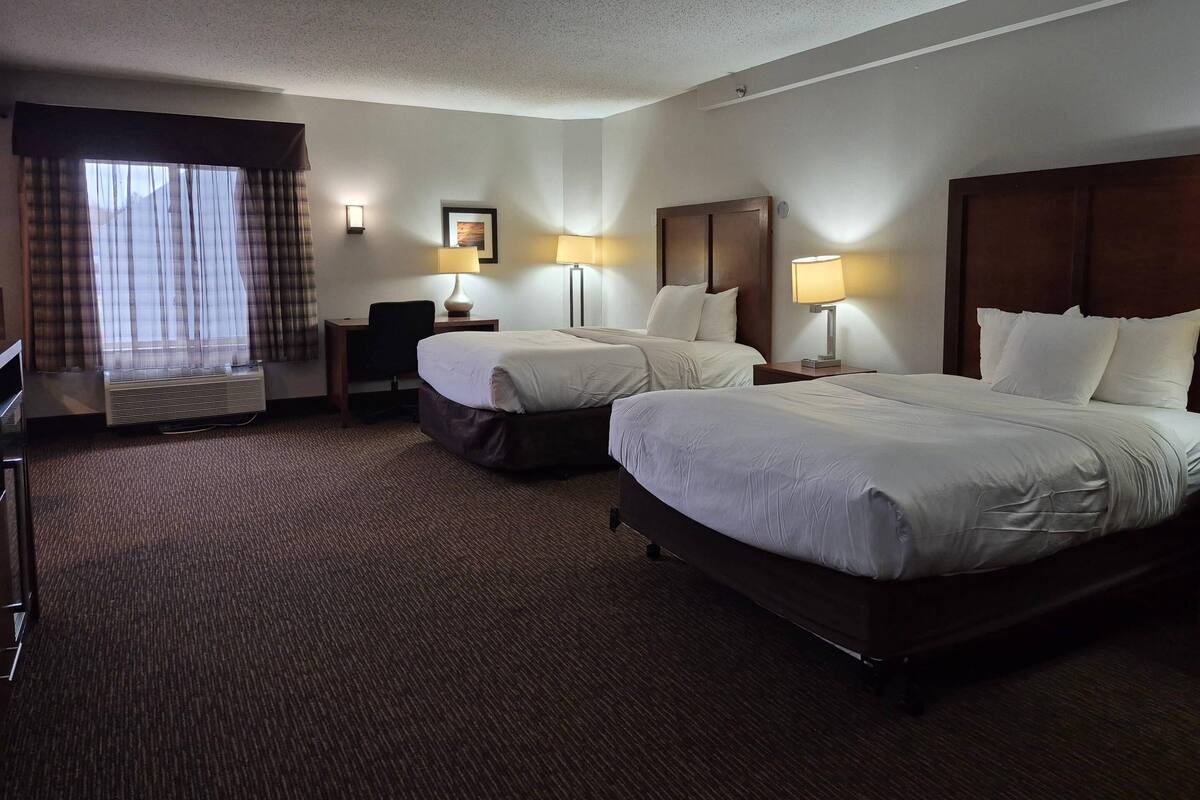 Quarto de hotel com duas camas, uma escrivaninha, uma cadeira, uma luminária de mesa e uma janela com cortinas.