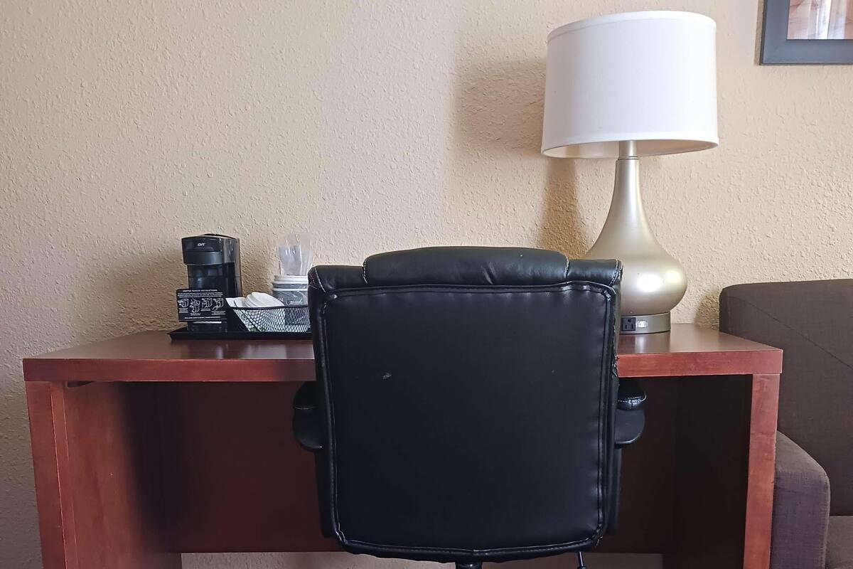 Quarto de hotel com escrivaninha de madeira, cadeira de escritório preta, luminária e sofá.