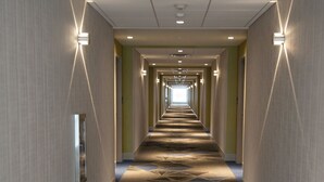 A long, narrow hotel corridor with patterned carpet, recessed lighting, and a mirrored wall on one side.