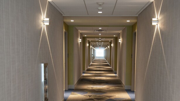 A long, narrow hotel corridor with patterned carpet, recessed lighting, and a mirrored wall on one side.