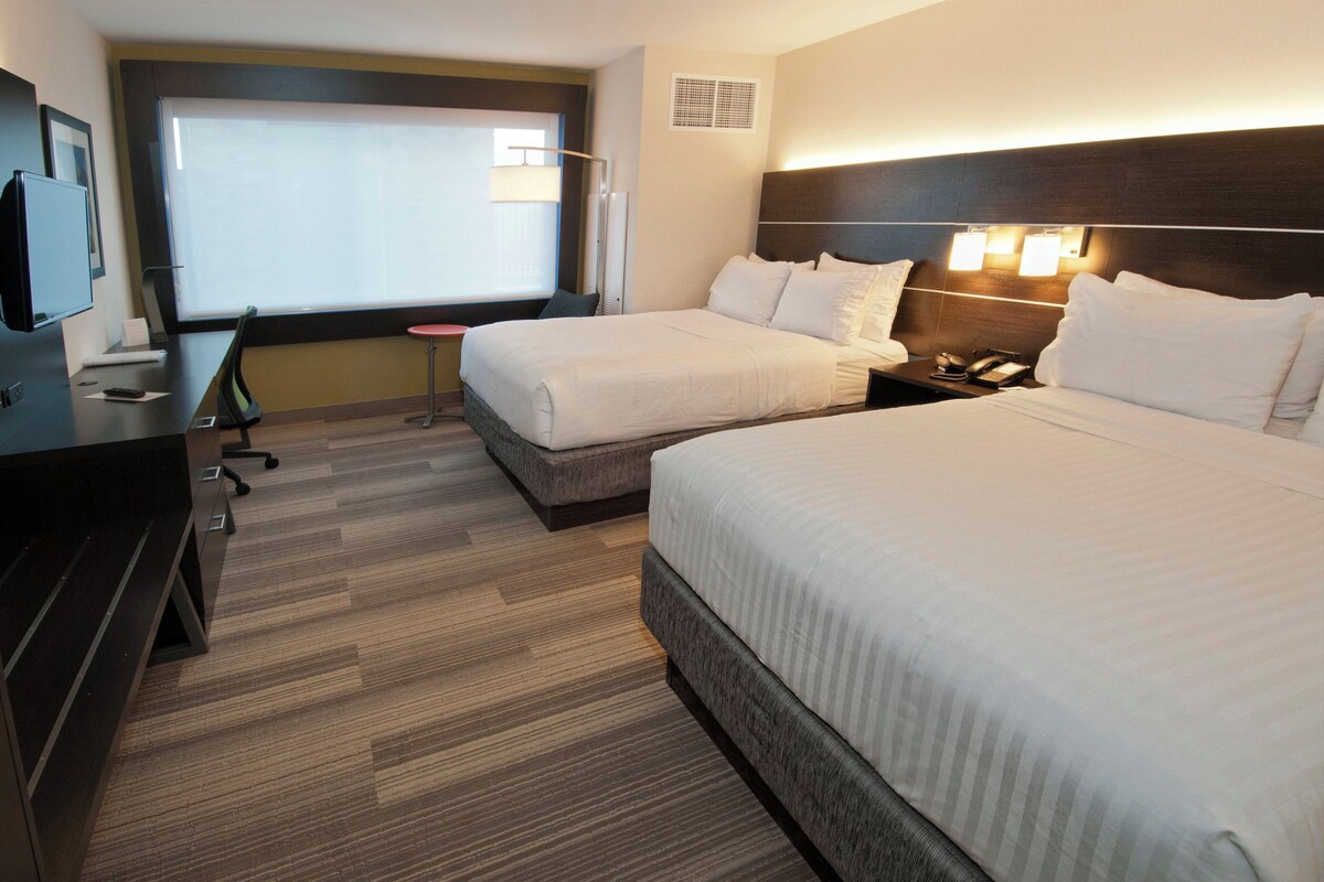 A hotel room with two beds, a desk, a chair, a TV, and a window with blinds.