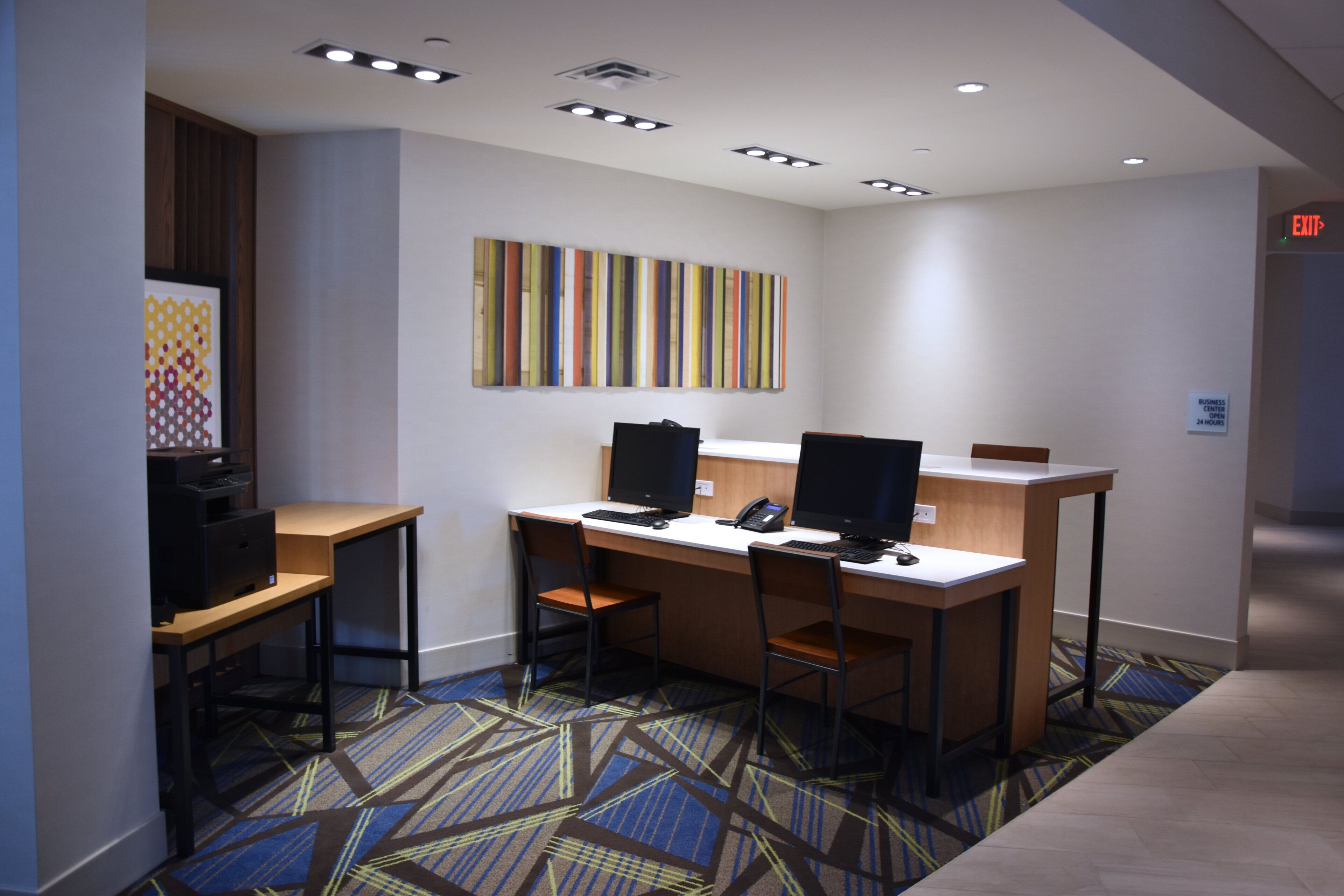 Un espace de travail aménagé dans une chambre d’hôtel, comprenant deux bureaux, des chaises, des ordinateurs et une œuvre d’art accrochée au mur.