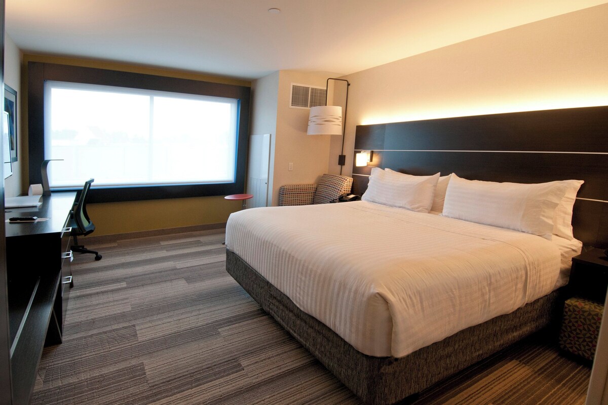 A hotel room with a large bed, a desk, a chair, a window, and a wall-mounted lamp.