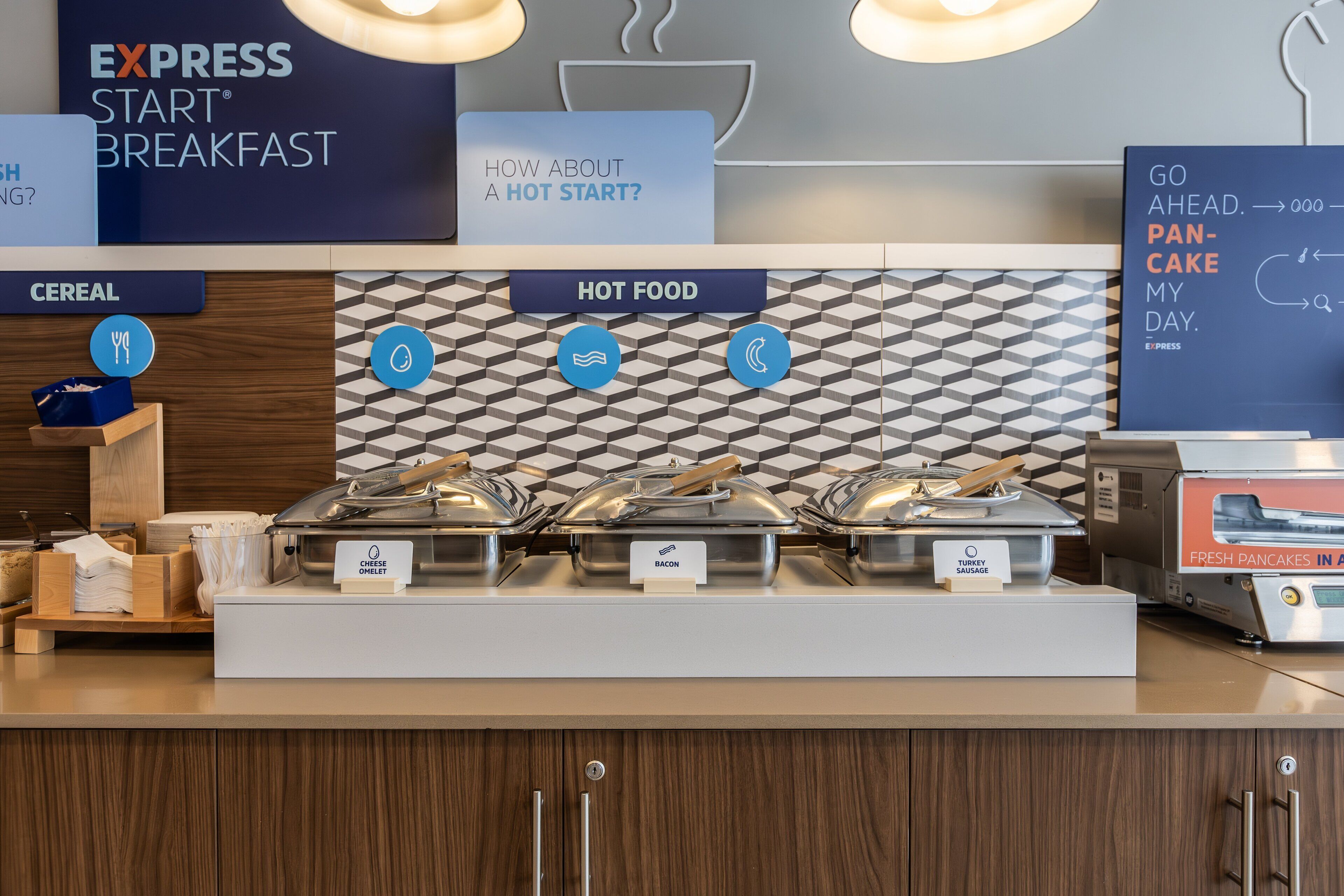 A buffet area with labeled sections for cereal and hot food, including cheese, bacon, sausage, and pancakes.