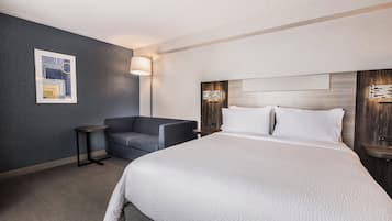 A hotel room with a large bed, a seating area with a chair, a wall-mounted lamp, and a framed picture.