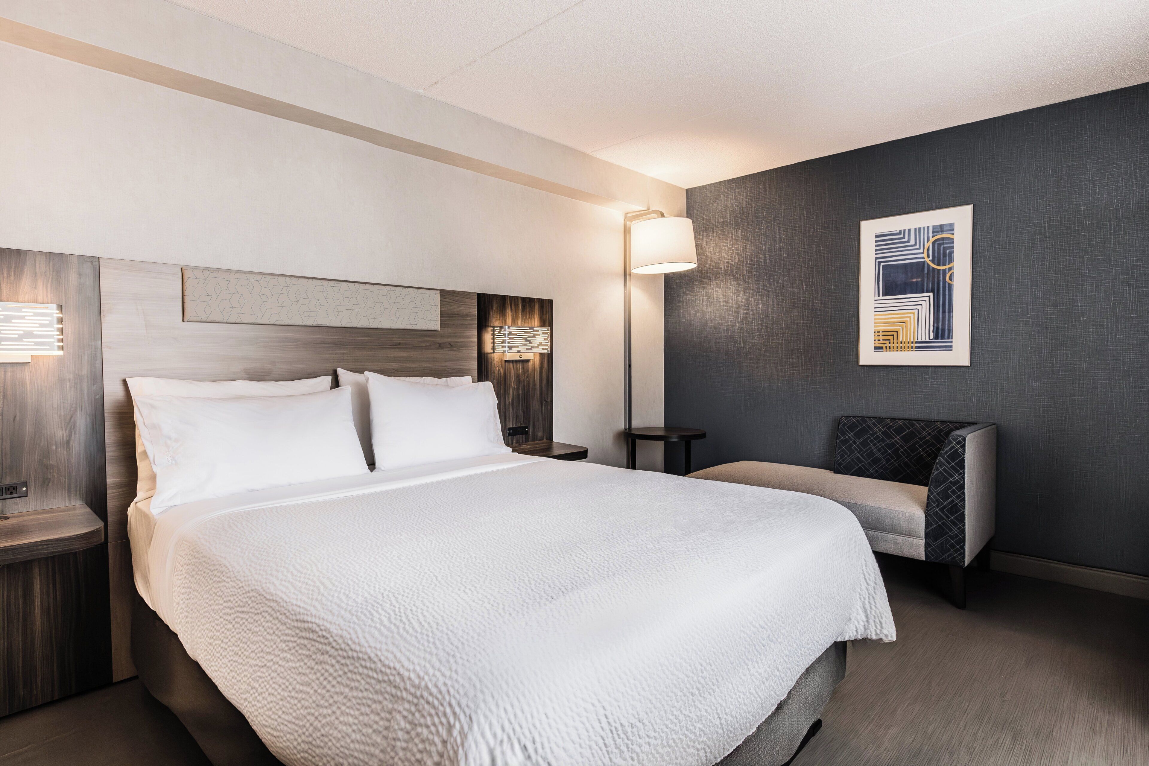 A hotel room with a large bed, bedside tables, a lamp, a chair, and a wall art piece.