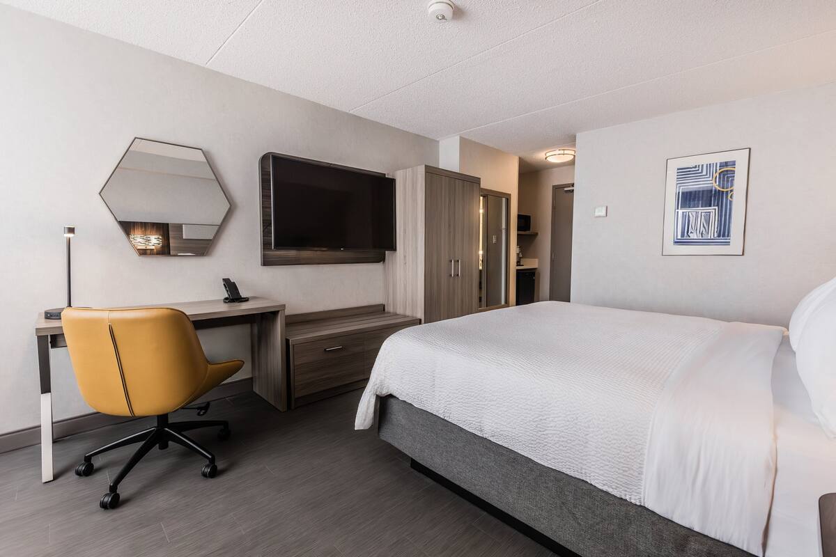 A hotel room with a bed, a desk, a chair, a wardrobe, a flat-screen TV, and a framed artwork on the wall.