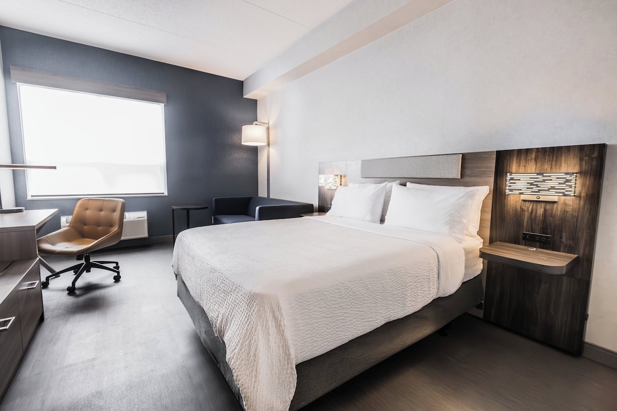 A modern hotel room with a large bed, a desk, a chair, and a nightstand.