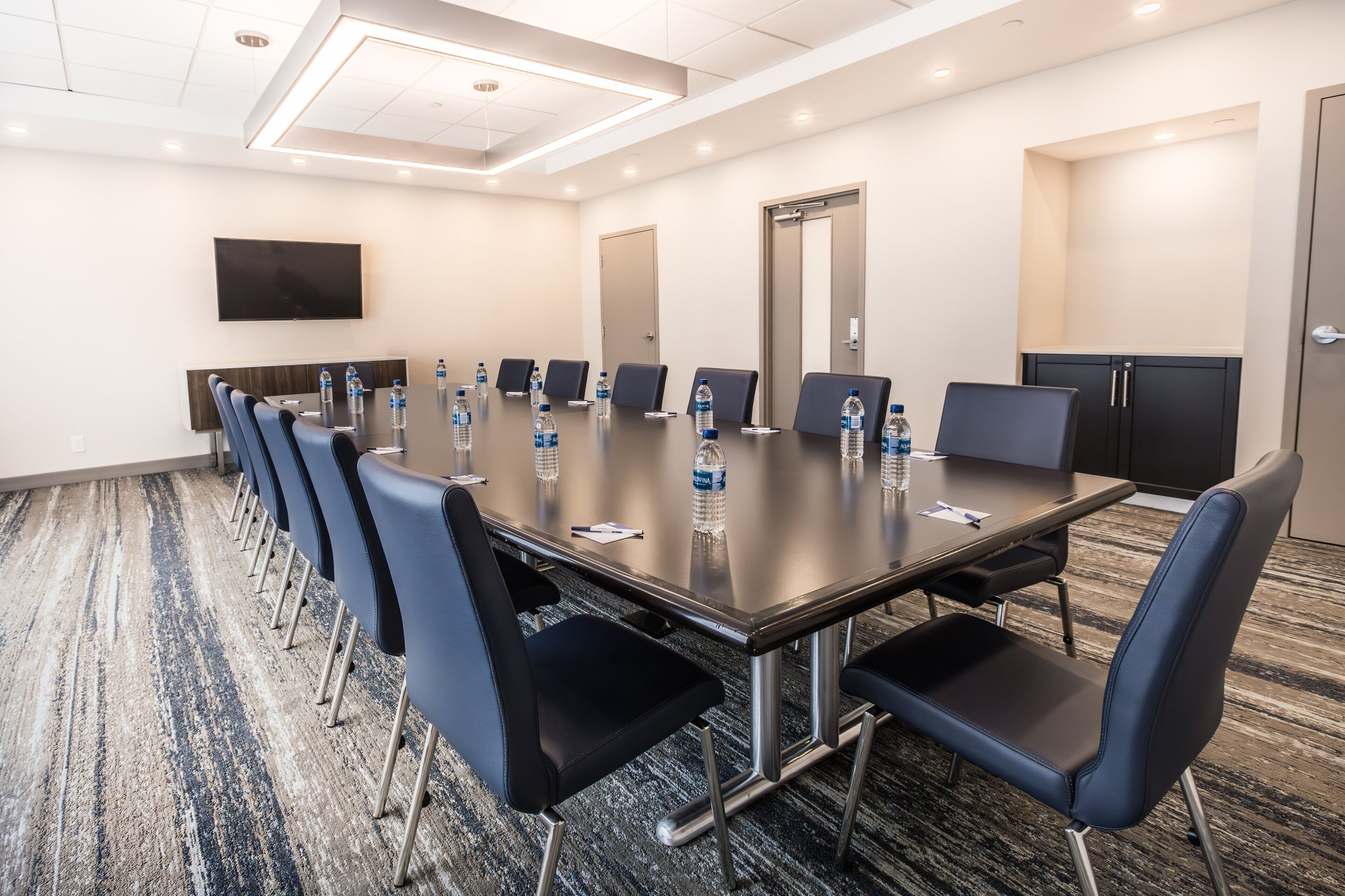 A conference room with a long table, blue chairs, water bottles, a flat-screen TV, and a carpeted floor.