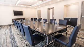 Une salle de conférence avec une longue table, des chaises bleues, des bouteilles d’eau, un téléviseur à écran plat et un sol recouvert de moquette.