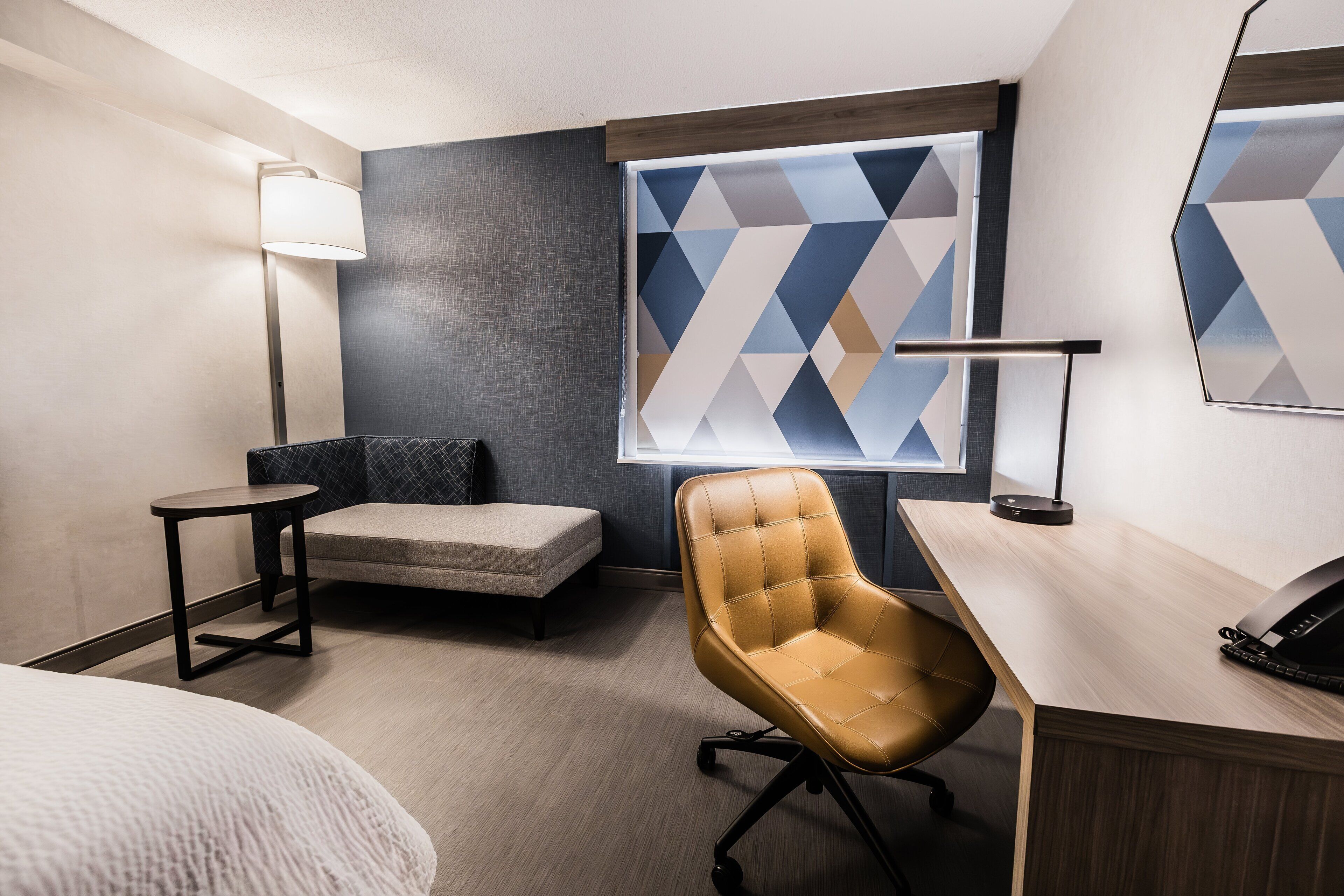 A hotel room with a bed, a desk with a telephone, a chair, a small table, a lamp, and a geometric-patterned window.
