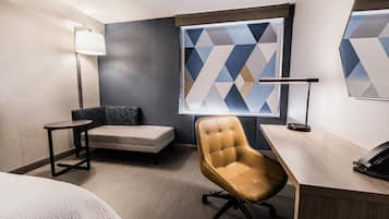 A hotel room with a bed, a desk with a telephone, a chair, a small table, a lamp, and a geometric-patterned window.