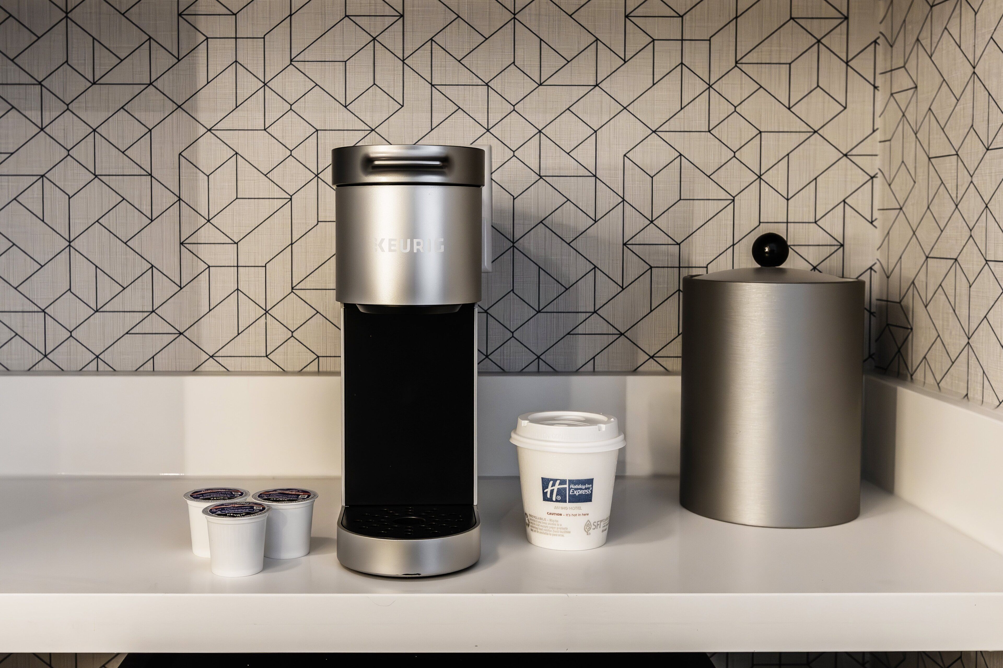 Cozinha moderna com uma cafeteira elegante, uma xícara de café e um recipiente cilíndrico em uma bancada branca.