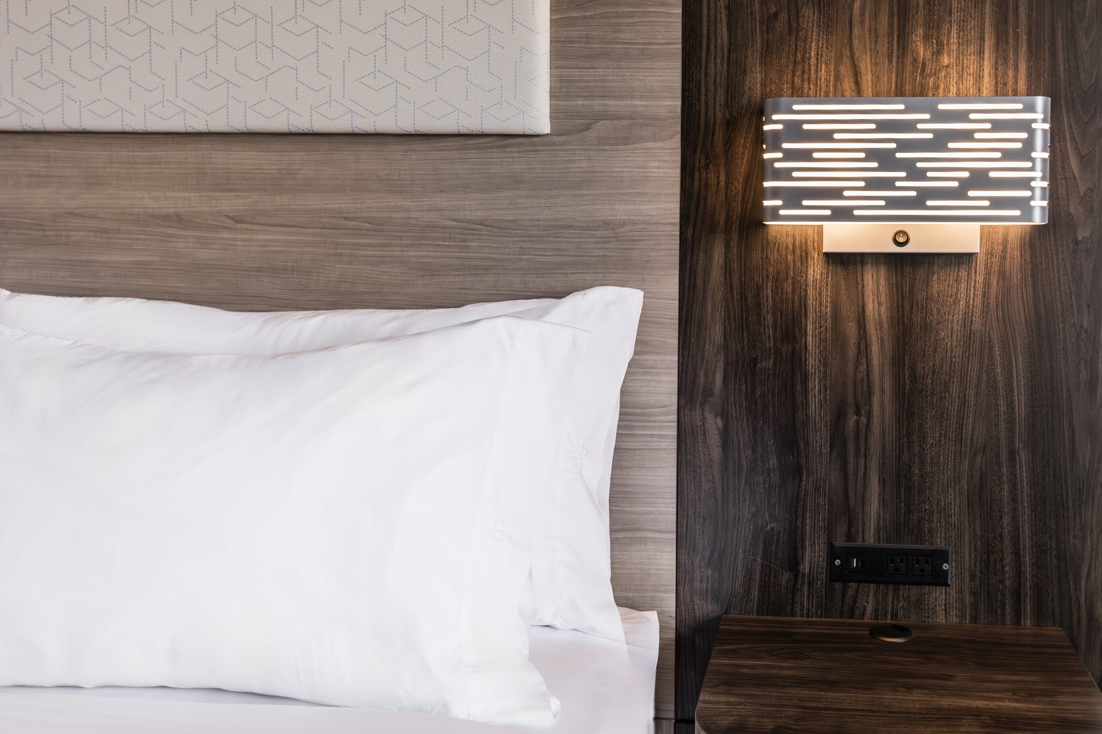Un letto rifatto con lenzuola bianche, una testiera in legno e un lampadario a parete con linee orizzontali.