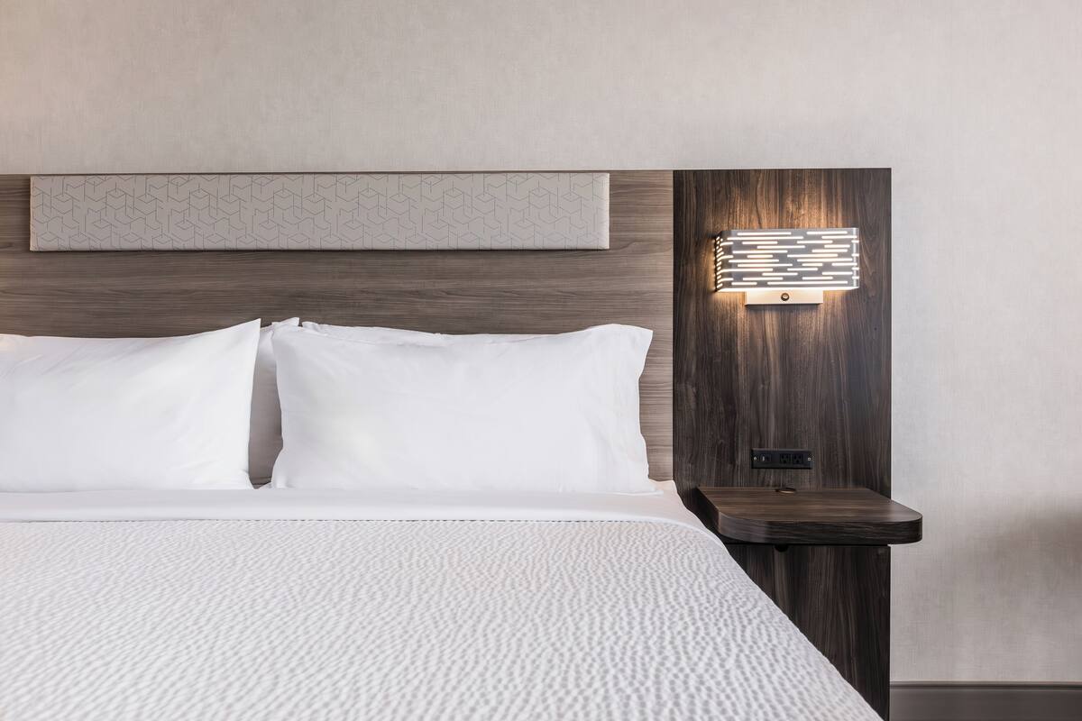 A modern hotel room with a large bed, a headboard with a geometric light panel, and a bedside table with a power outlet.