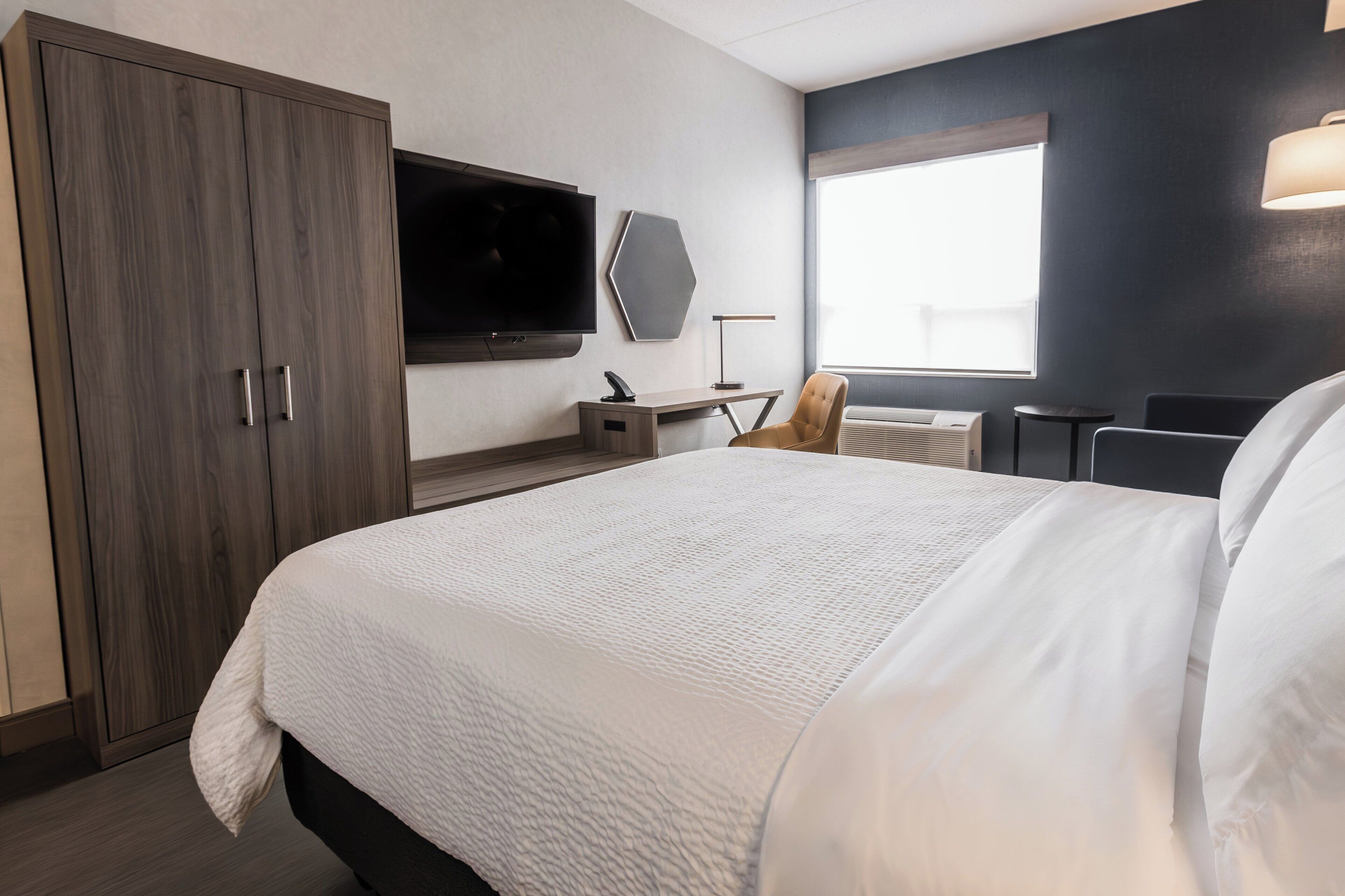 Ein Hotelzimmer mit einem großen Bett, einem flachbildfernseher an der Wand, einem Schreibtisch mit Stuhl und einem Fenster, das für natürliches Licht sorgt.