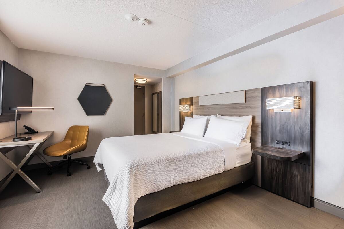 A modern hotel room with a large bed, a desk with a chair, a TV, and a wall-mounted light fixture.
