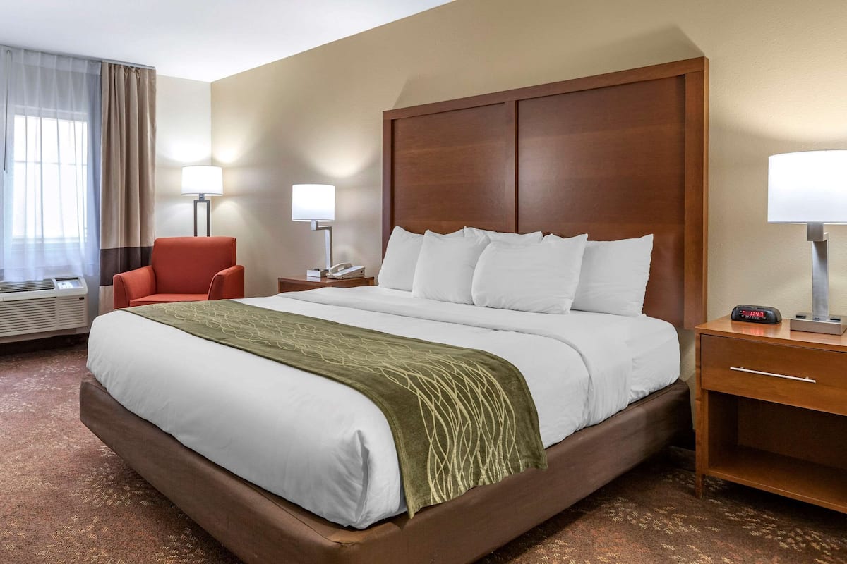 A hotel room with a large bed, a wooden headboard, two bedside tables with lamps, a red armchair, and a window with curtains.