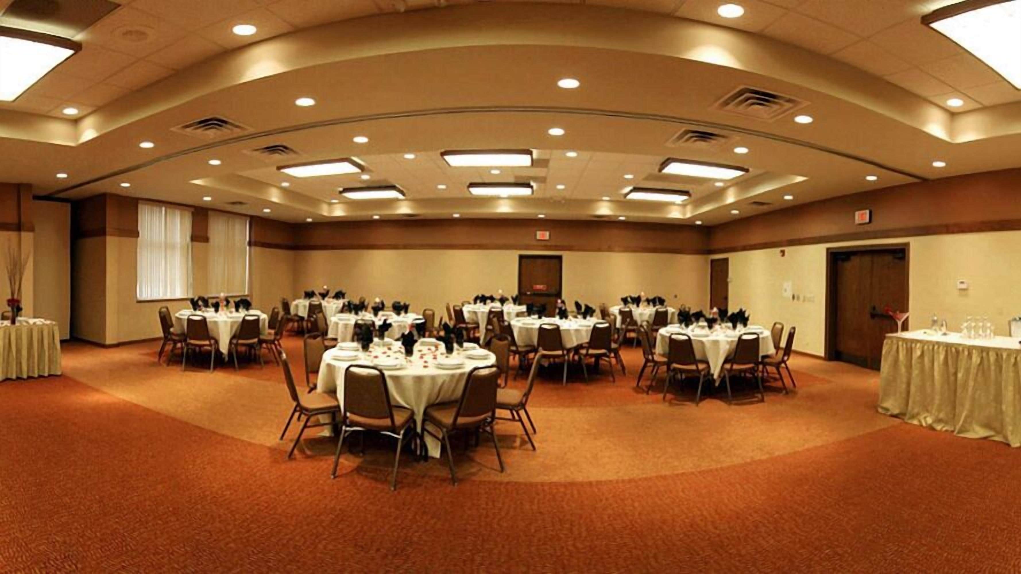 A large conference room with multiple round tables, each set with white tablecloths and chairs, and a carpeted floor.