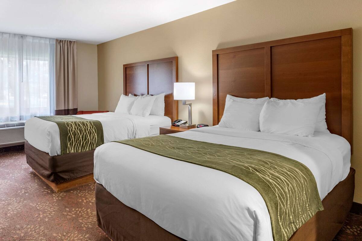 A hotel room with two beds, a nightstand with a lamp, and a window with curtains.
