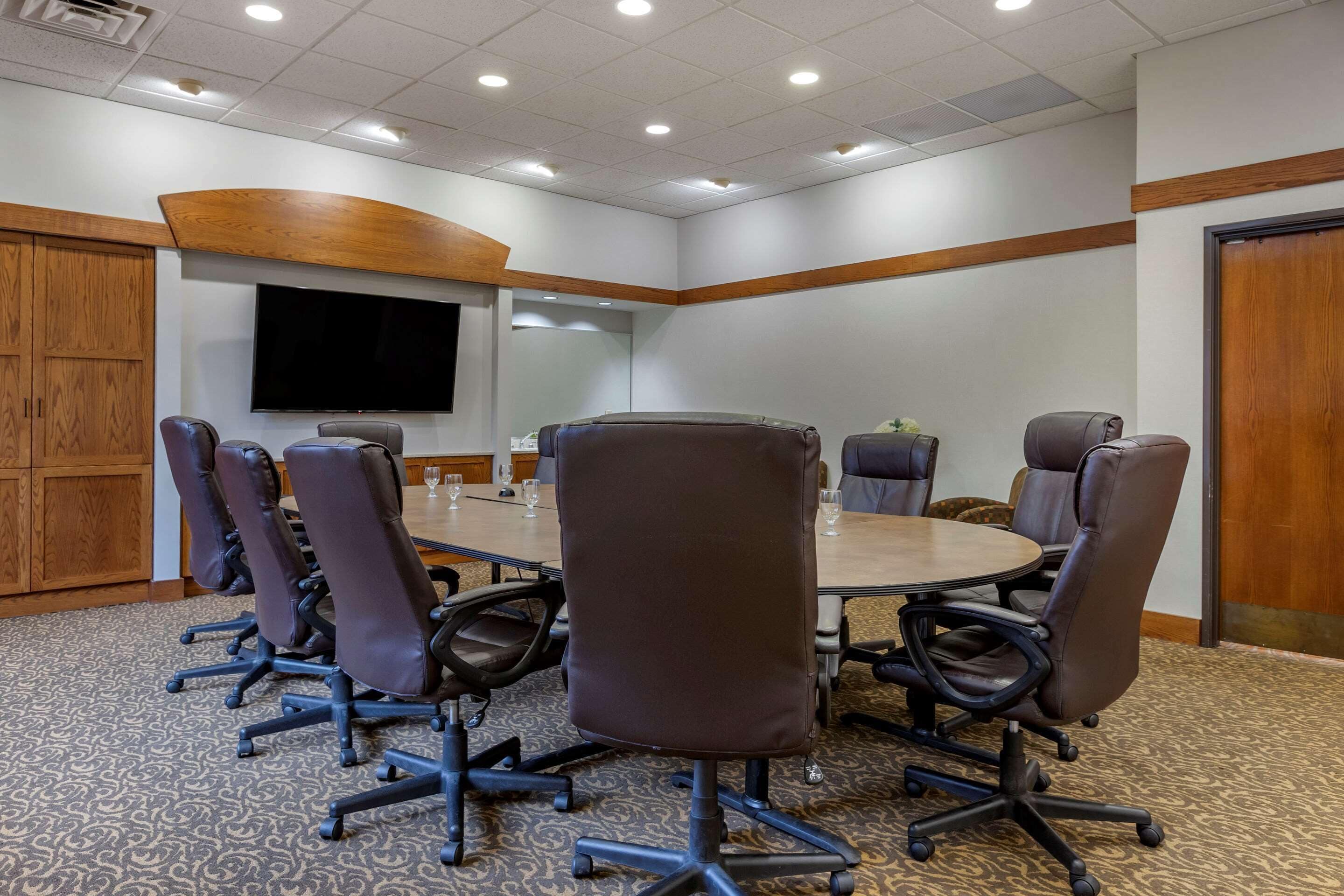 Een vergaderzaal met een grote tafel, meerdere stoelen, een flatscreen televisie, houten lambrisering en een kamerbreed tapijt met dessin.