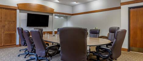 Een vergaderzaal met een grote tafel, meerdere stoelen, een flatscreen televisie, houten lambrisering en een kamerbreed tapijt met dessin.