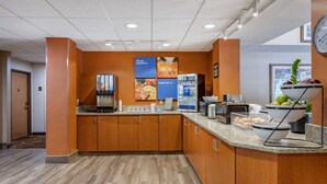 Een moderne keukenhoek met een self-service ontbijtstation, inclusief een koffieapparaat, een koelkast en een schaal met fruit.