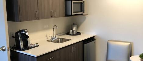 A compact kitchen with dark wood cabinets, a microwave, a coffee maker, and a sink. There is a trash can and a small table with a chair.